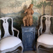 Styled vignette showing the carved wood figure displayed atop a blue-painted cabinet between two upholstered armchairs