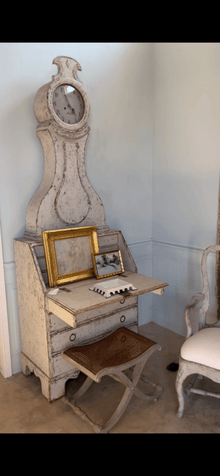 Caned stool styled in front of an antique painted Mora clock secretary desk with gilt-framed prints on the fold-down top