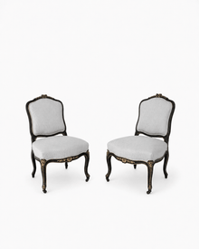 Pair of ebonized Louis XV-style side chairs with gilt floral accents, cabriole legs, and gray striped upholstery, shown on a white background