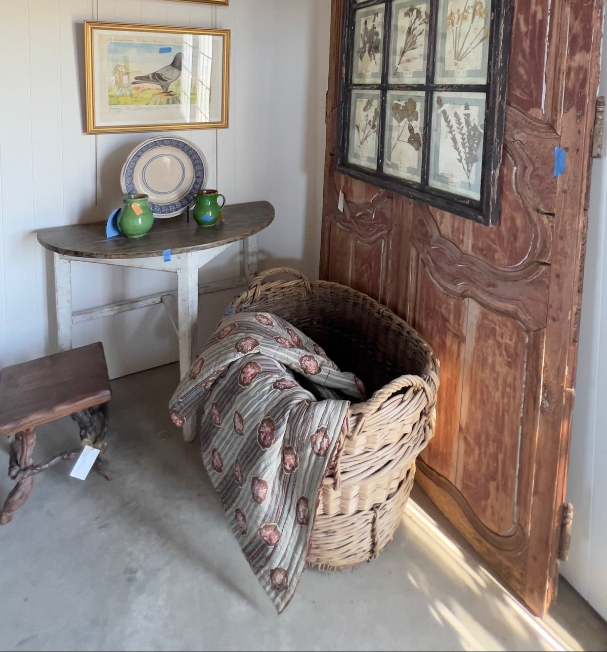 Shop vignette showing the framed pigeon print hung above a demilune table with ceramics, near a woven basket and antique door