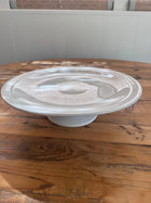 Single low-profile white ironstone cakestand shown alone on a wood table, showing its shallow rimmed plate and short pedestal