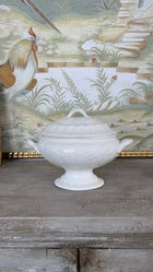 The tureen displayed on a weathered wood surface in front of a framed painted rooster and garden scene